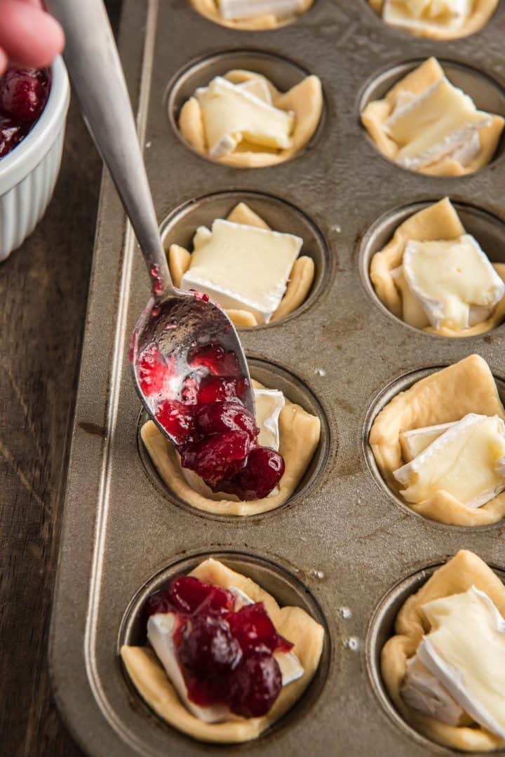 a spoon putting cranberry sauce on top of brie in muffin pan.