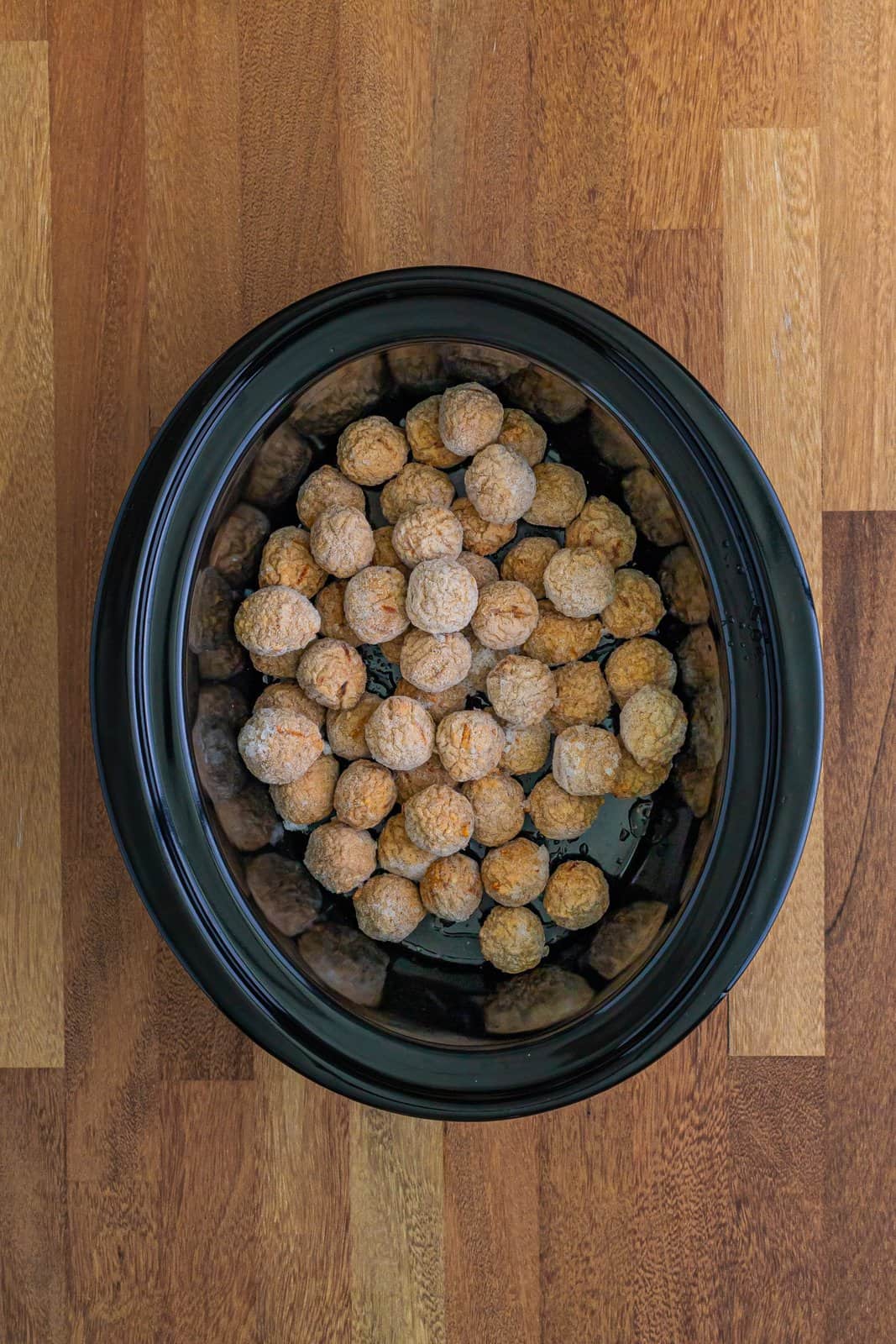 frozen meatballs shown at the bottom of an oval black slow cooker. 