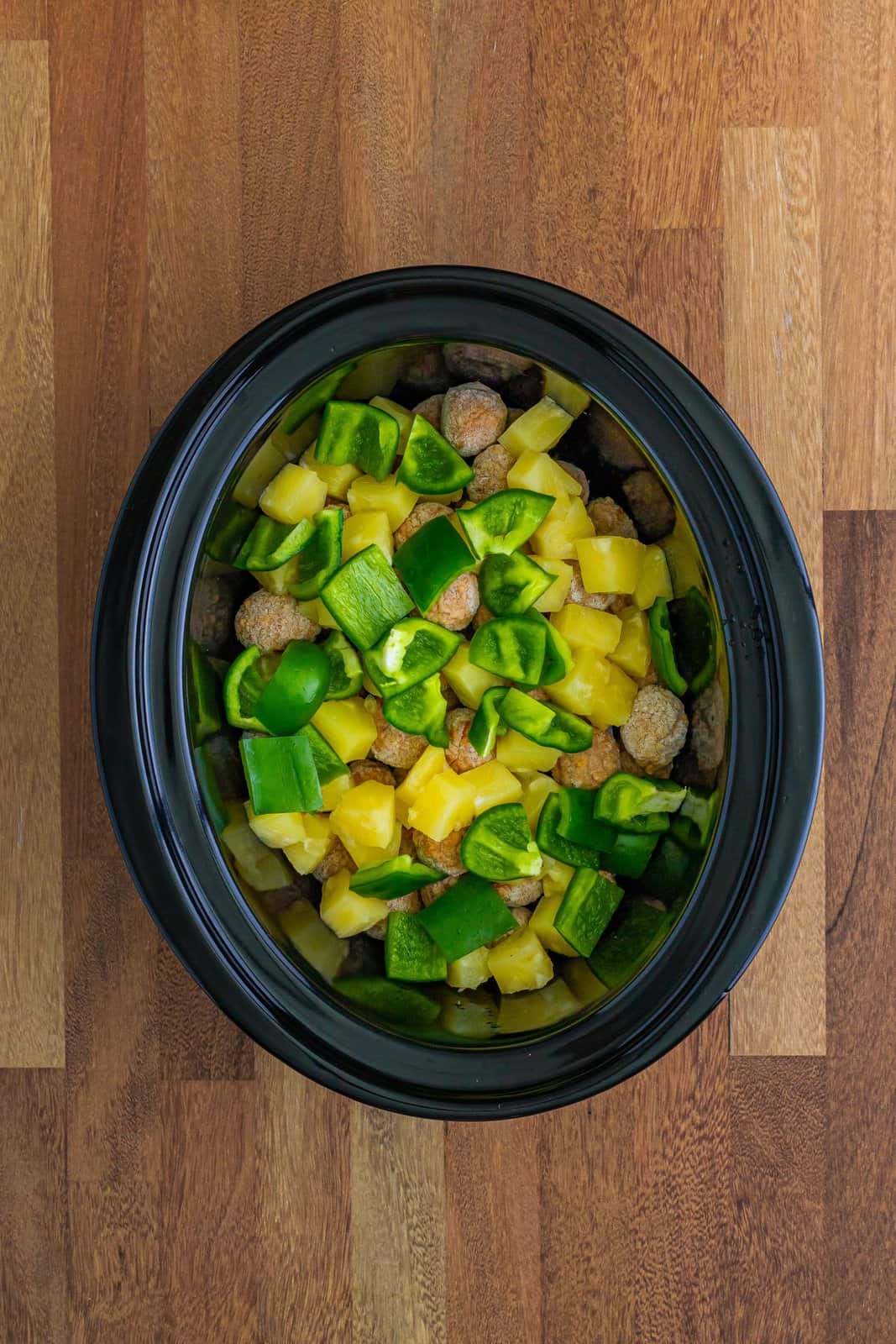 chopped green peppers and pineapple chunks shown on top of the frozen meatballs in a slow cooker. 