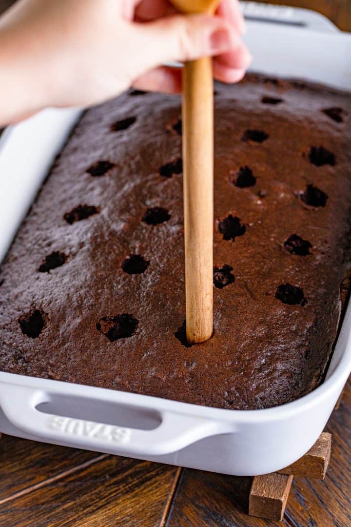 a wooden spoon handle being shown poking holes into baked cake. 