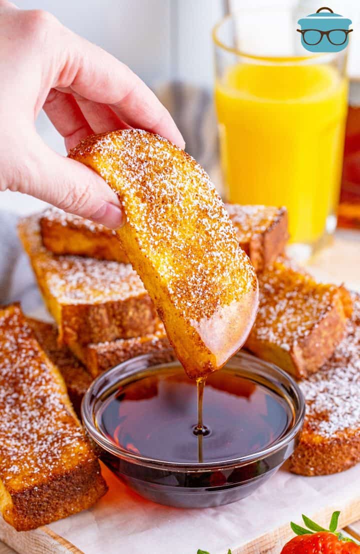 a hand dipping a French toast stick into maple syrup.
