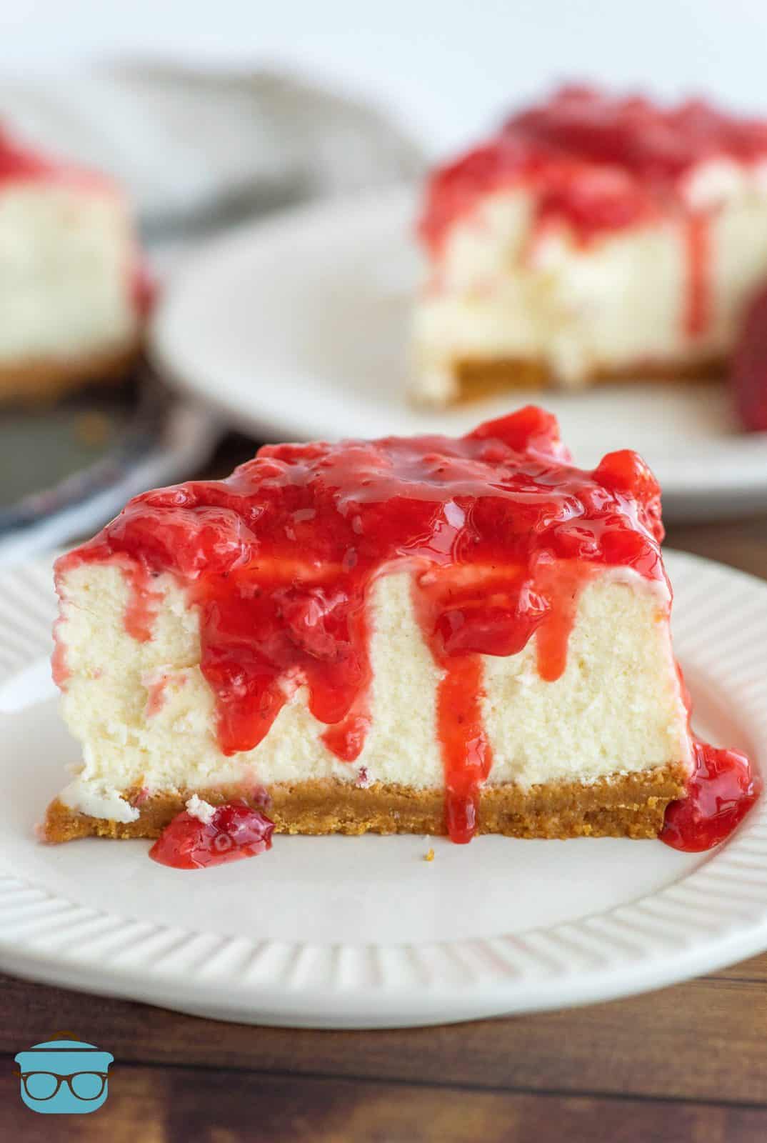 Slice of New York Cheesecake on white plate with strawberry sauce over top.