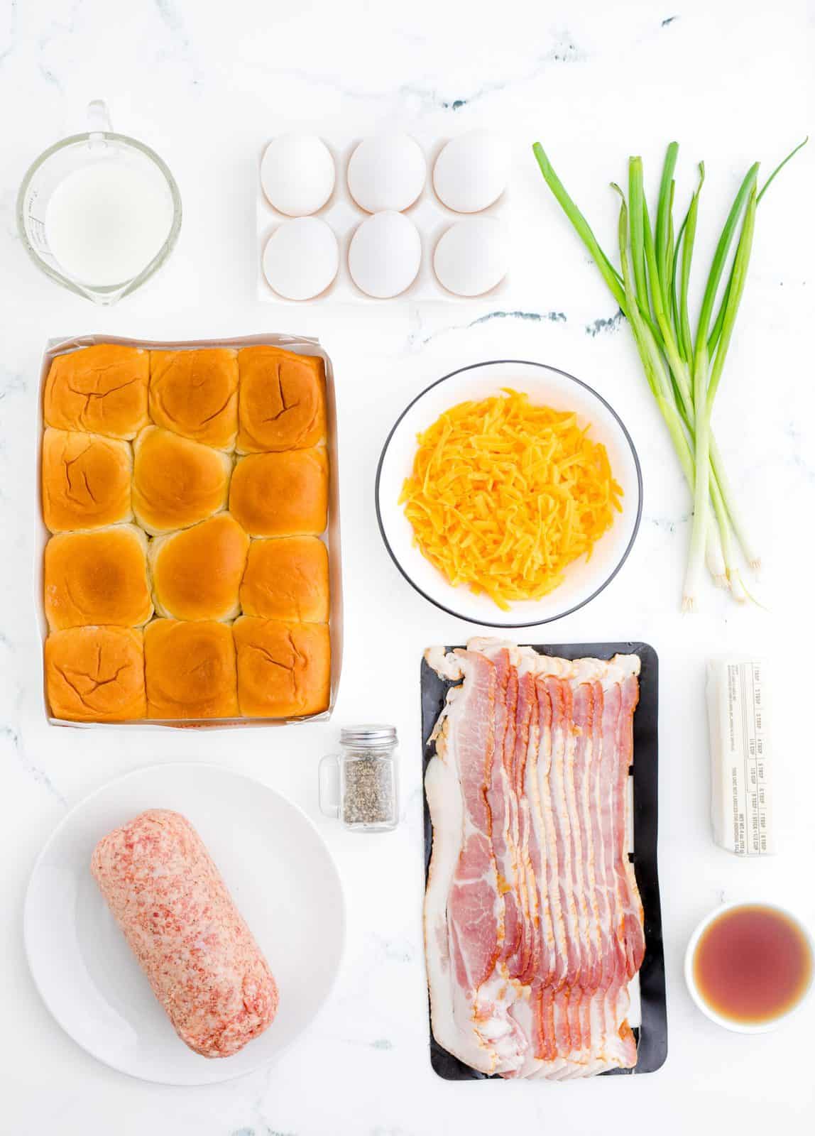 Ingredients needed for breakfast sliders: pork sausage, bacon, eggs, milk, chives, pepper, dinner rolls, shredded cheddar cheese, salted butter and maple syrup.