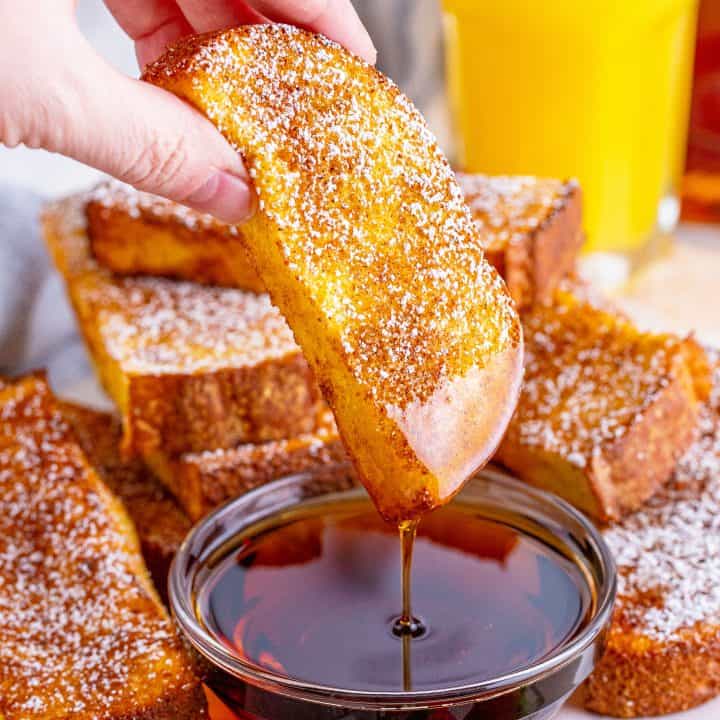 A hand dipping a French toast stick into a small bowl of maple syrup.