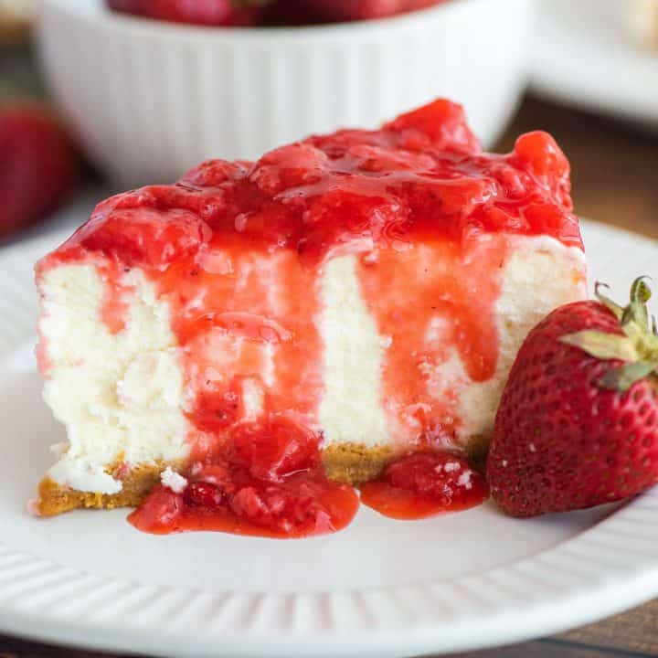 Square image of slice of cheesecake with sauce dripping down side with strawberry on plate.