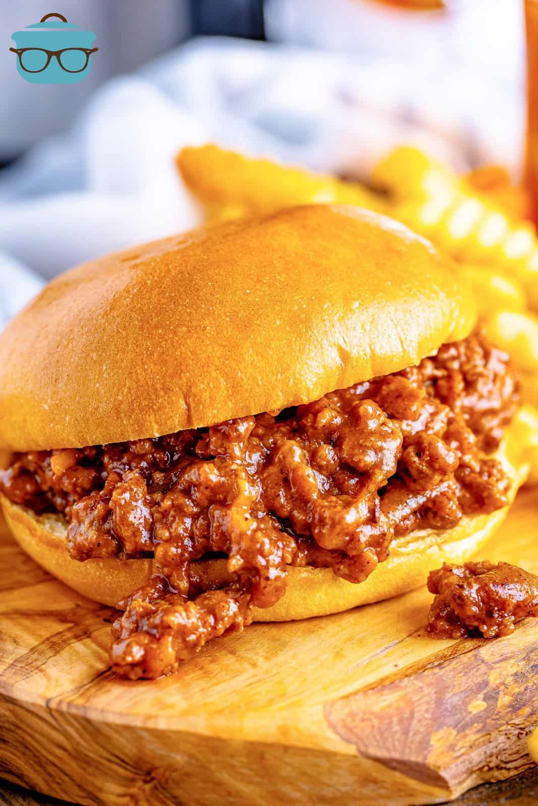 a sloppy Joe sandwich on a wood surface with French fries in the background. 