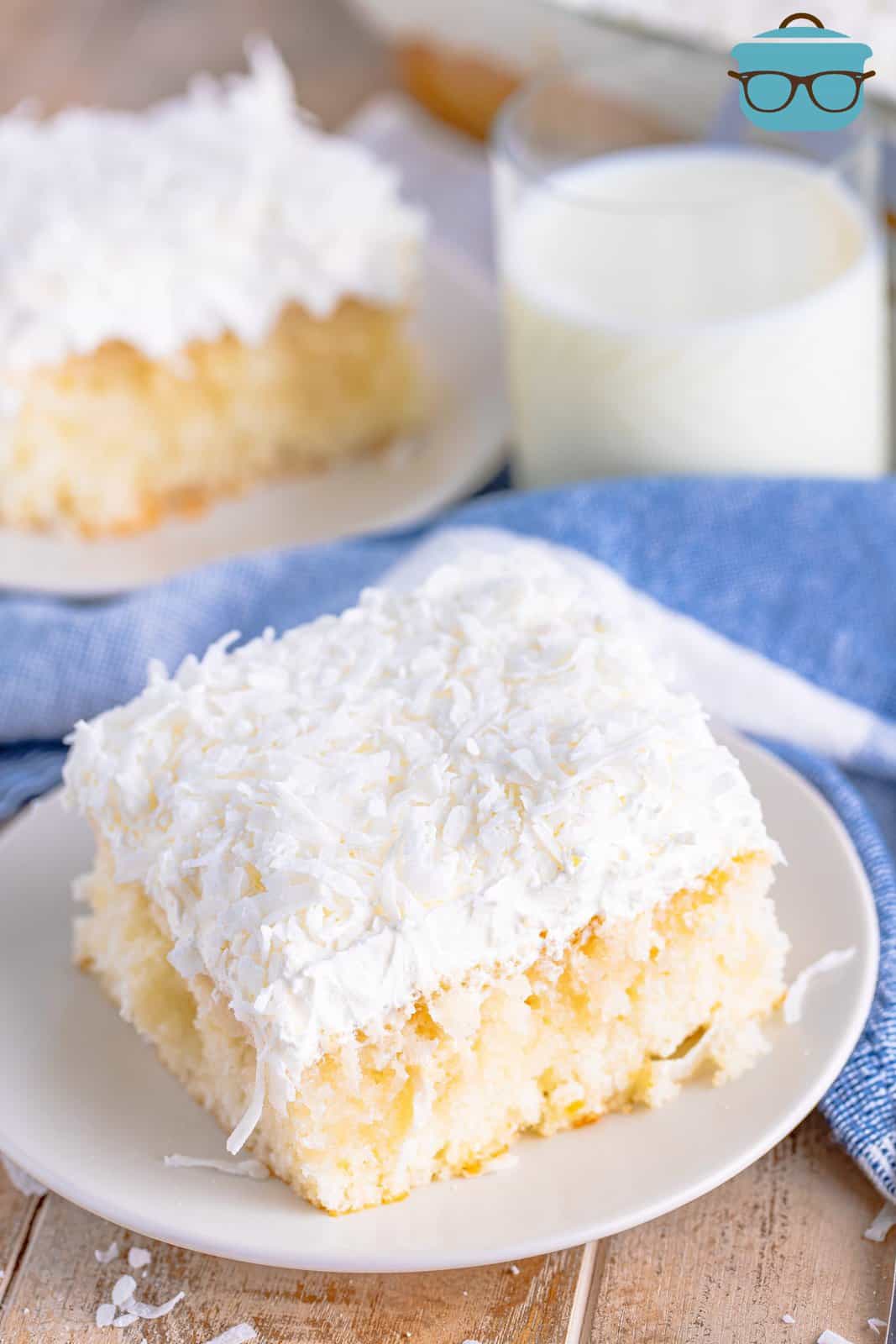 a slice of coconut cake on a small white plate with a glass of milk shown in the background.