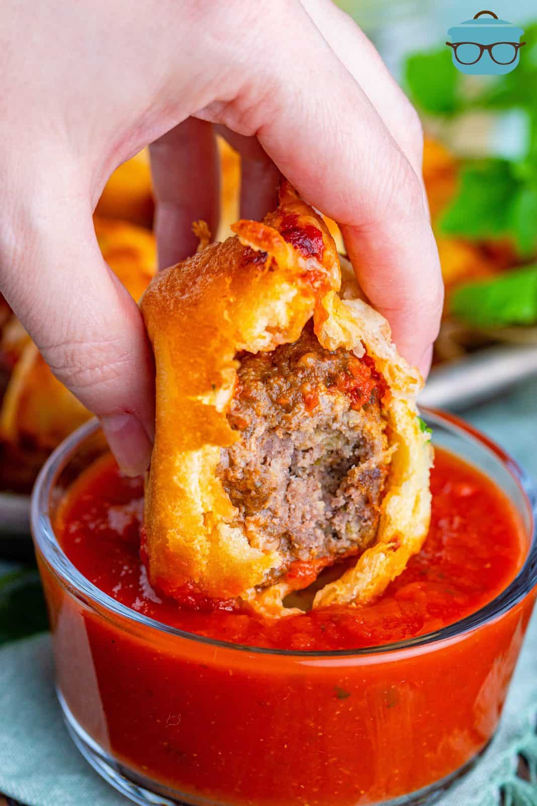 Hand dipping one Meatball Sub Cup in marinara sauce with bite taken out showing inside.