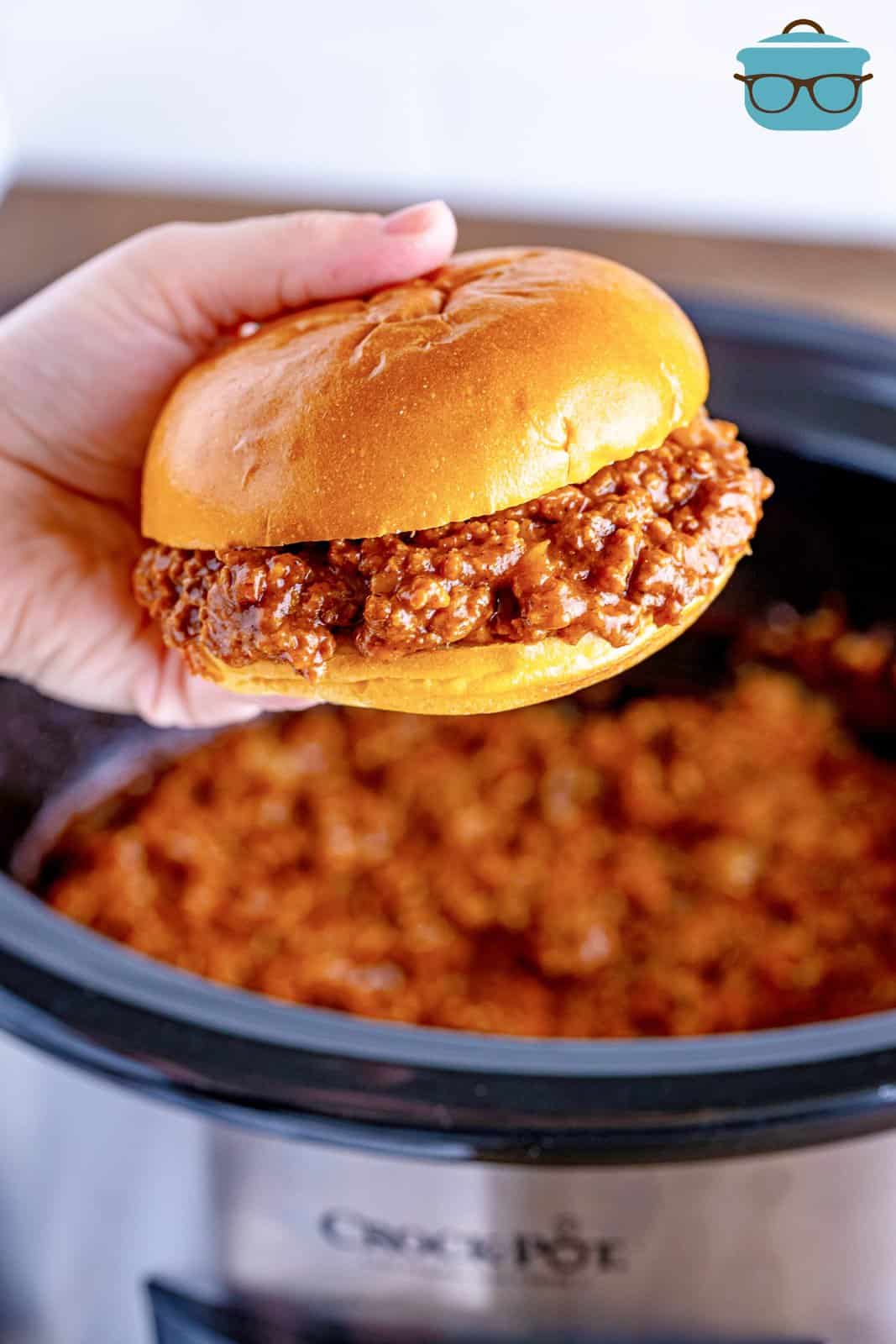 a hand holding a sloppy Joe over a slow cooker. 