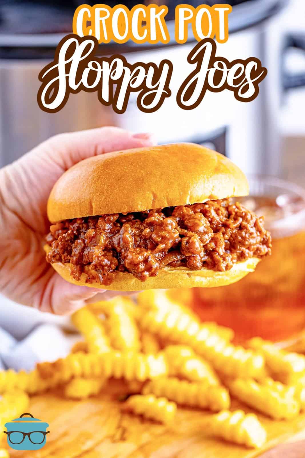 a hand holding up a Sloppy Joe sandwich over a plate of French fries with a glass of iced tea in the background.