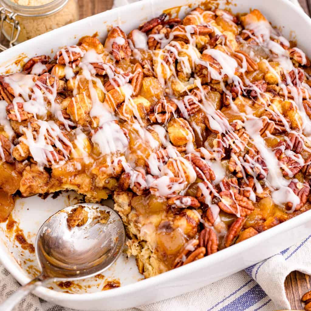 Square image of Cinnamon Roll Casserole in dish with some missing and serving spoon in dish.