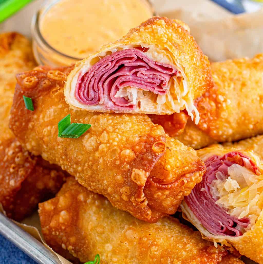 Square image of Reuben Egg Rolls stacked on tray with top one cut in half showing inside.