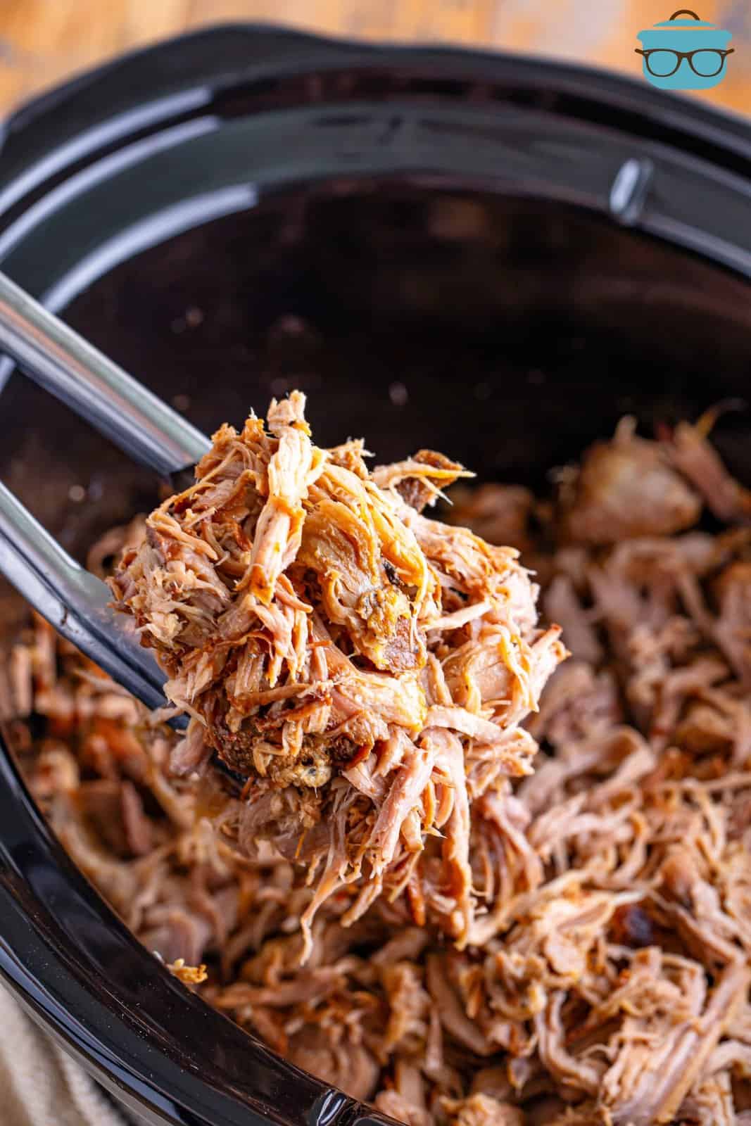 a pair of tongs holding up some pulled pork over the slow cooker. 