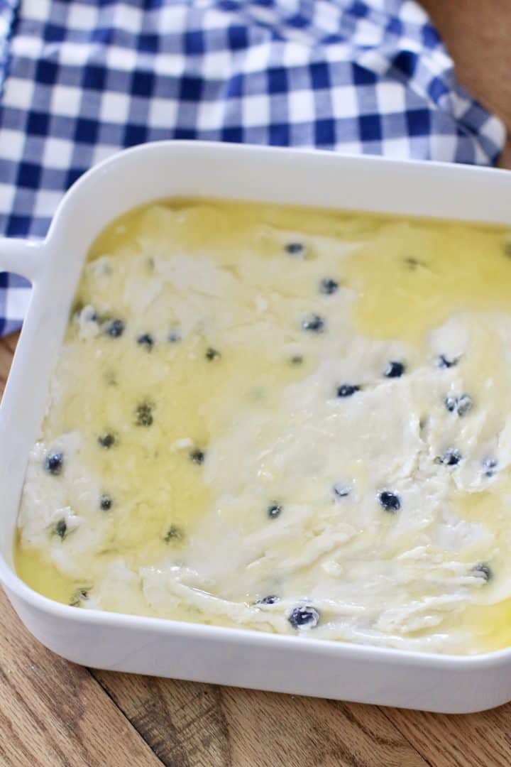blueberry biscuit dough spread into a white baking dish with melted butter.
