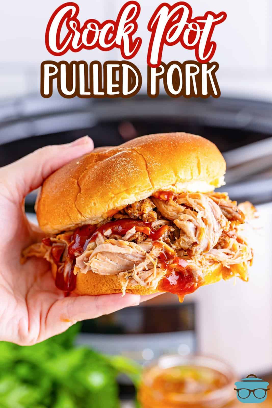 a hand holding up a pulled pork sandwich in front of a slow cooker. 