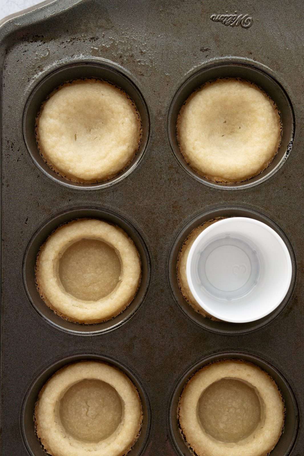Baked cookie dough out of oven with shot glass pressing down to reshape the cookie cups.