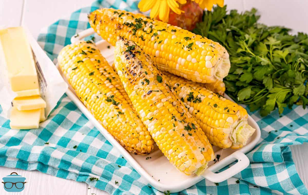 Rectangle image of Grilled Corn on white platter with butter and herbs.