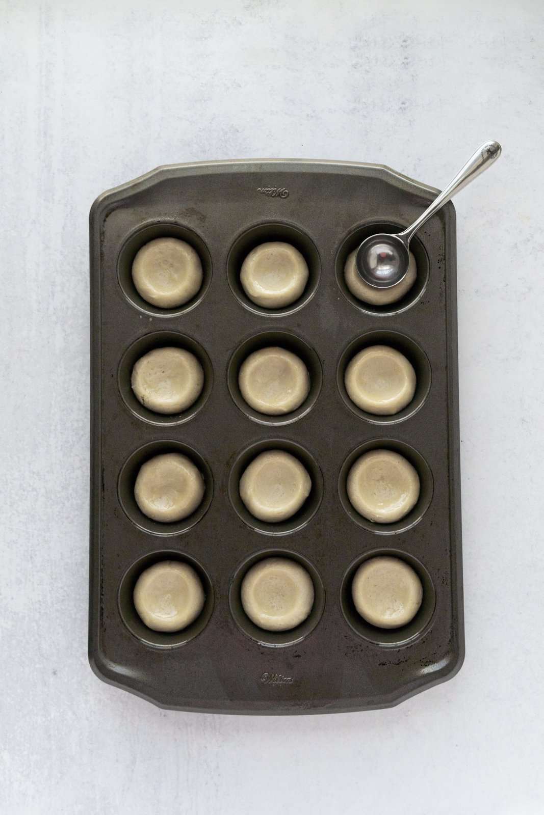 Cookie dough added to mini muffin tin with measuring spoon pressing down depressions in dough.