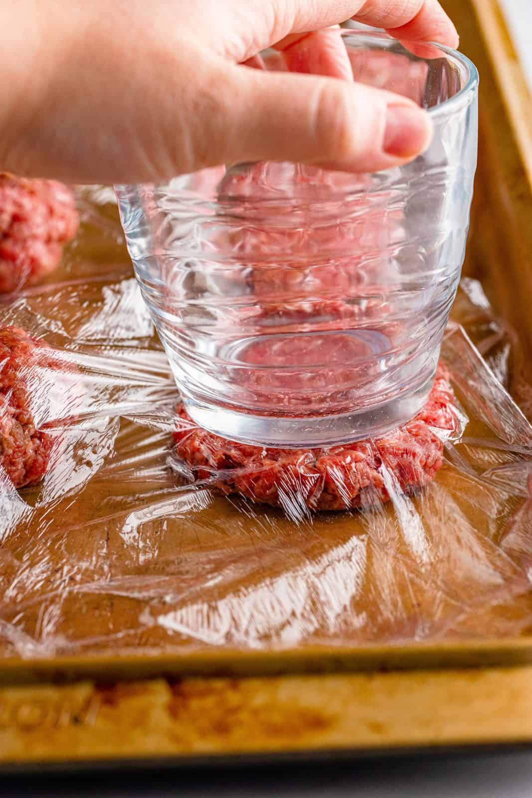 Glass pressing burger patties down with glass over plastic wrap.