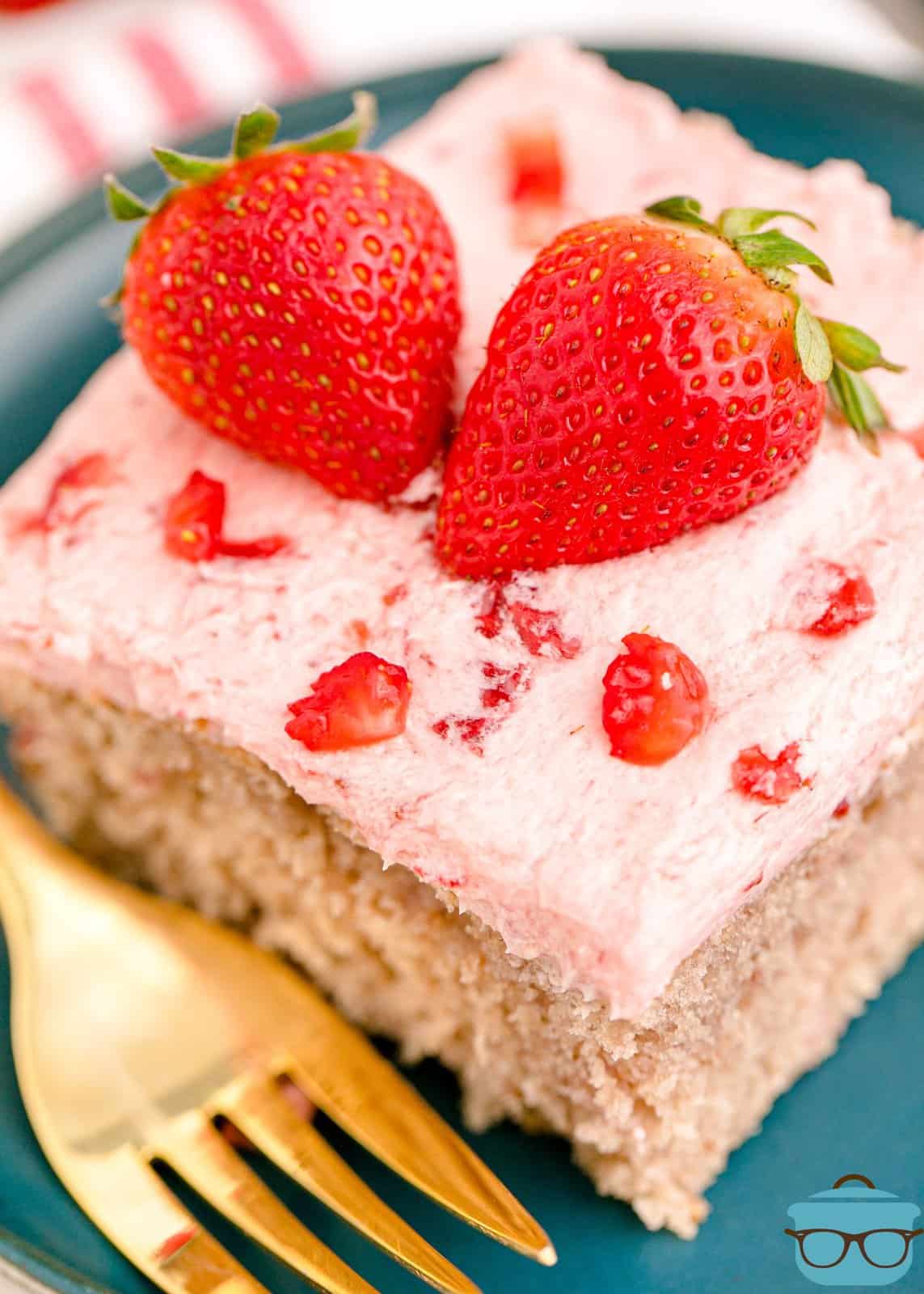 Overhead photo slice of Fresh Strawberry Sheet Cake on plate with gold fork topped with strawberry halves.