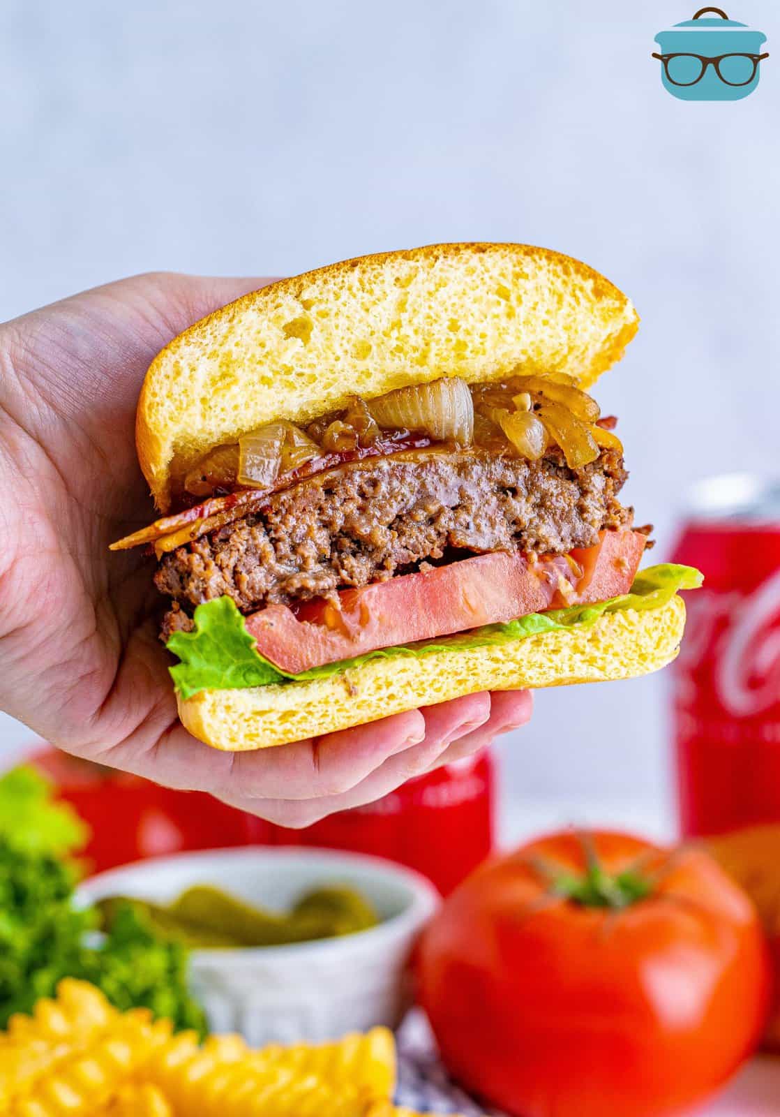 Hand holding up one of the Grilled Bacon Cheeseburgers with Coca Cola Onions sliced in half showing the inside.