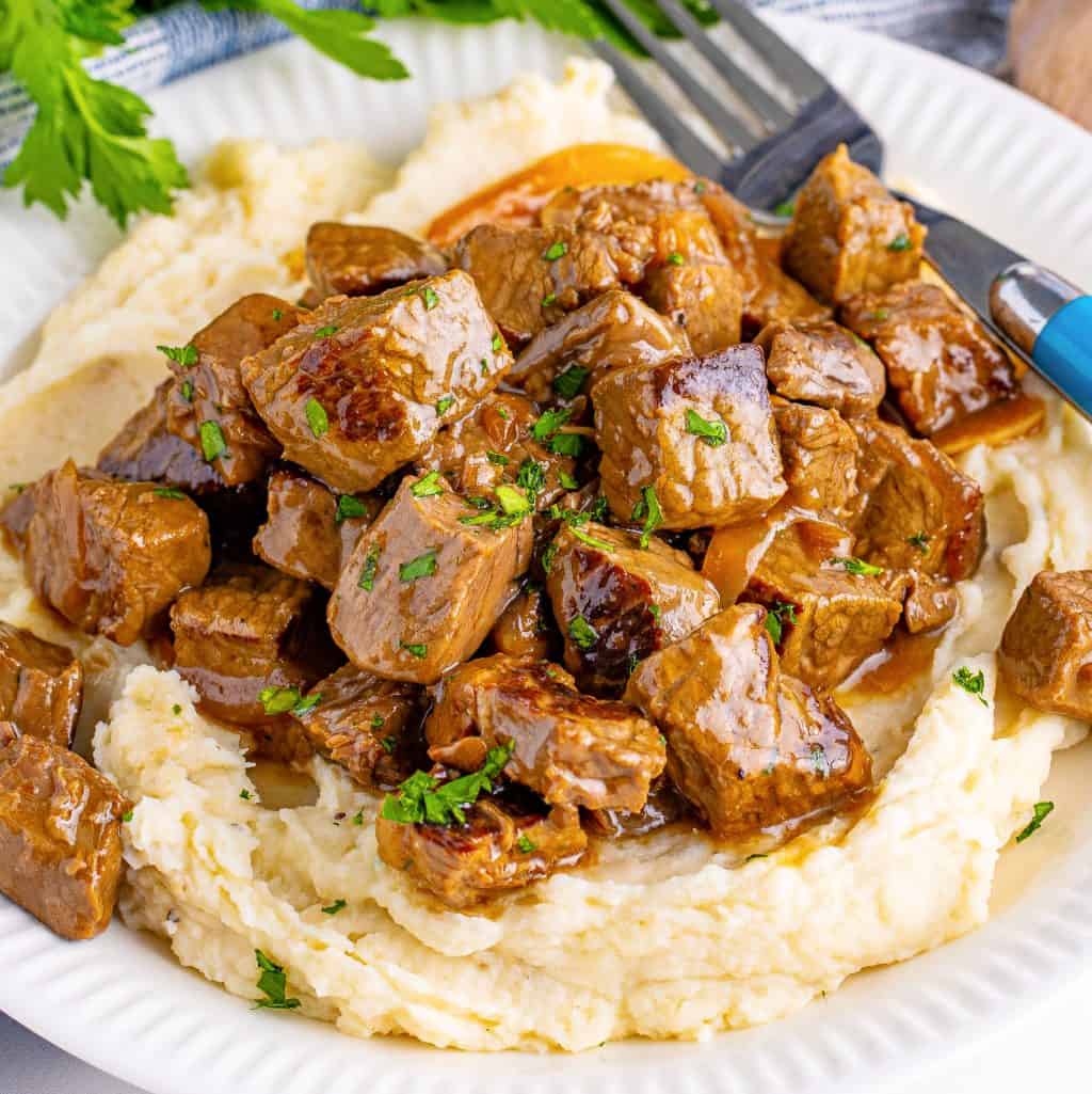 Slow Cooker Garlic Butter Steak Bites close up square image on plate with mashed potatoes and fork.