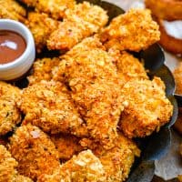 Square image of stacked Cornflake Chicken Nuggets on black platter with sauce.