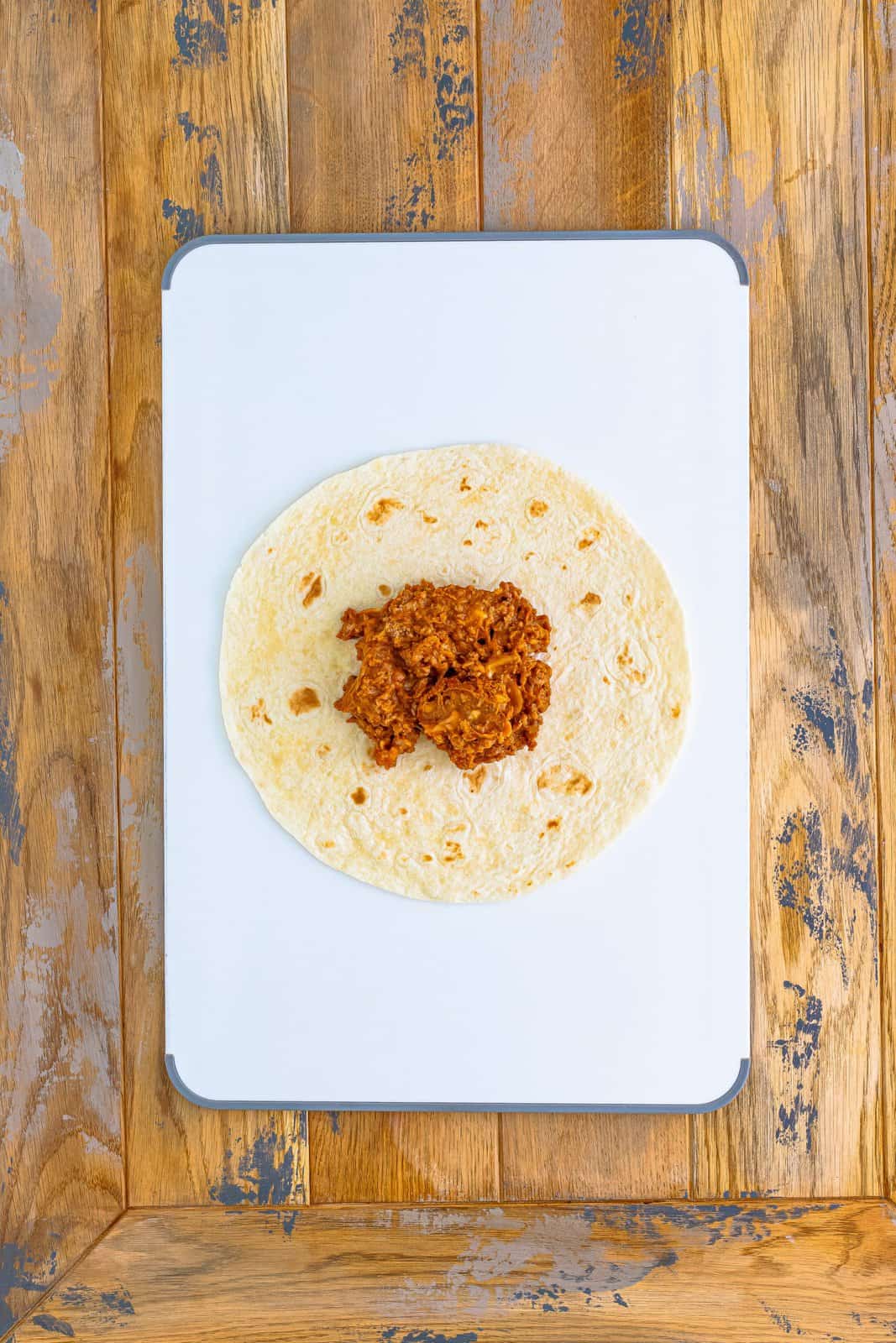 One tortilla on cutting board with filling placed in the center.