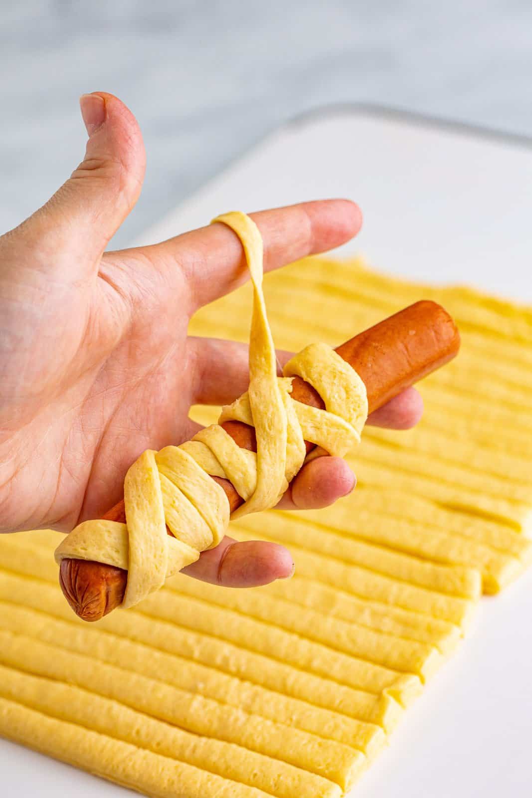 Hand wrapping hod dog in crescent dough strips.
