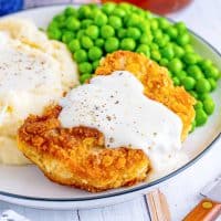 Close up square image of Country Fried Pork Chops and Gravy on white plate with mashed potatoes and peas.