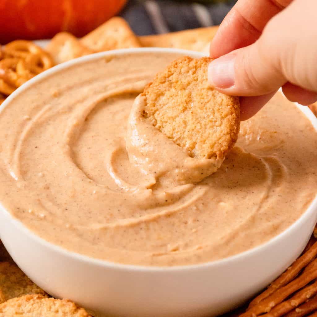 Square image close up of hand dipping a cookie into Pumpkin Dip in bowl.