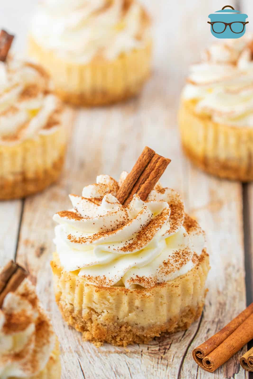 Egg Nog Mini Cheesecakes on white board topped with whipped cream, cinnamon and cinnamon stick.