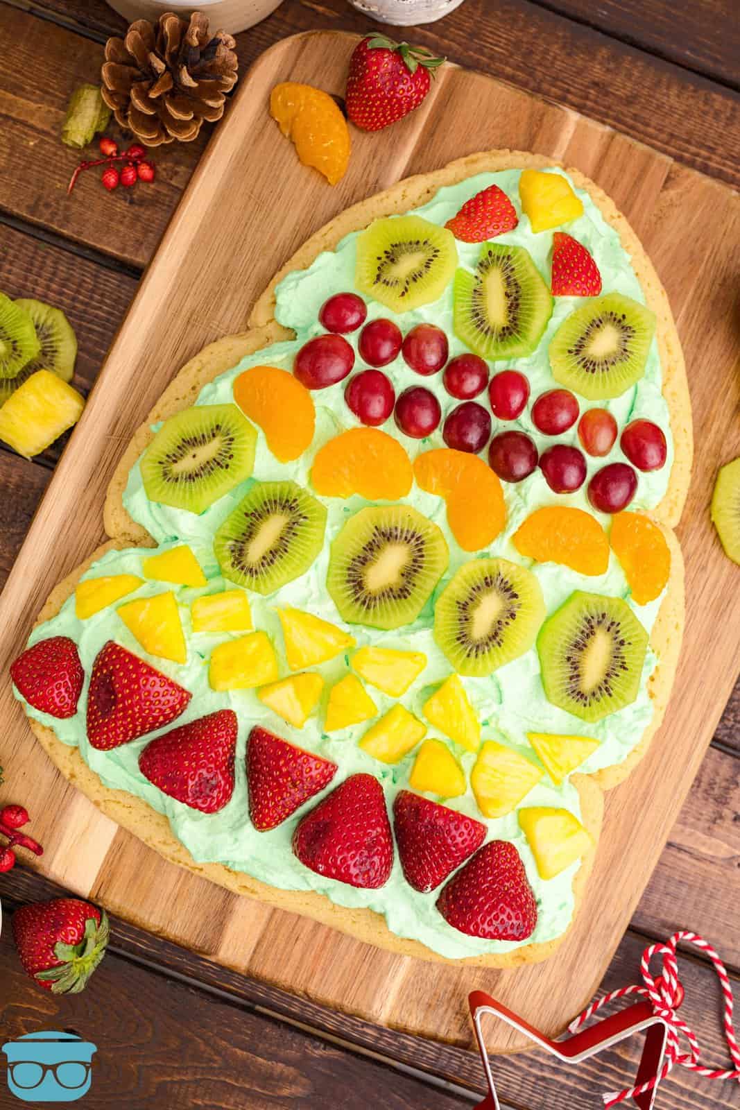Overhead of Christmas Tree Fruit Pizza on wooden cutting board with fruit around it.