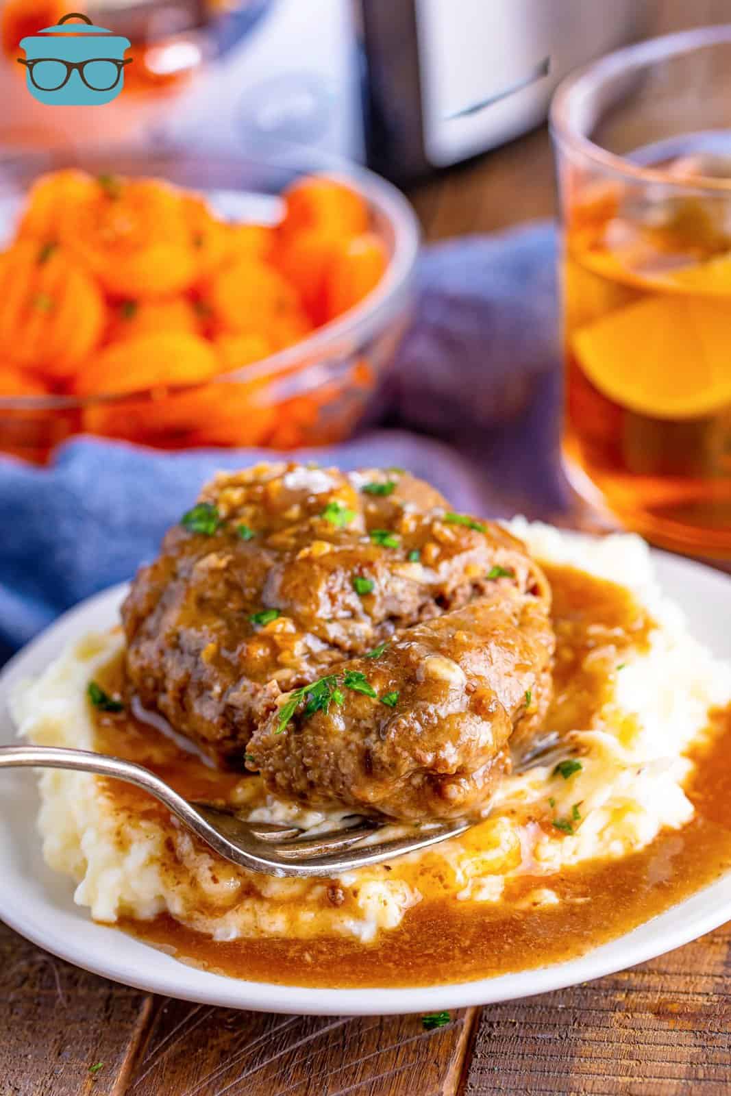 salisbury steak with gravy shown served on a a bed of mashed potatoes with a bowl of steamed sliced carrots in the background. 