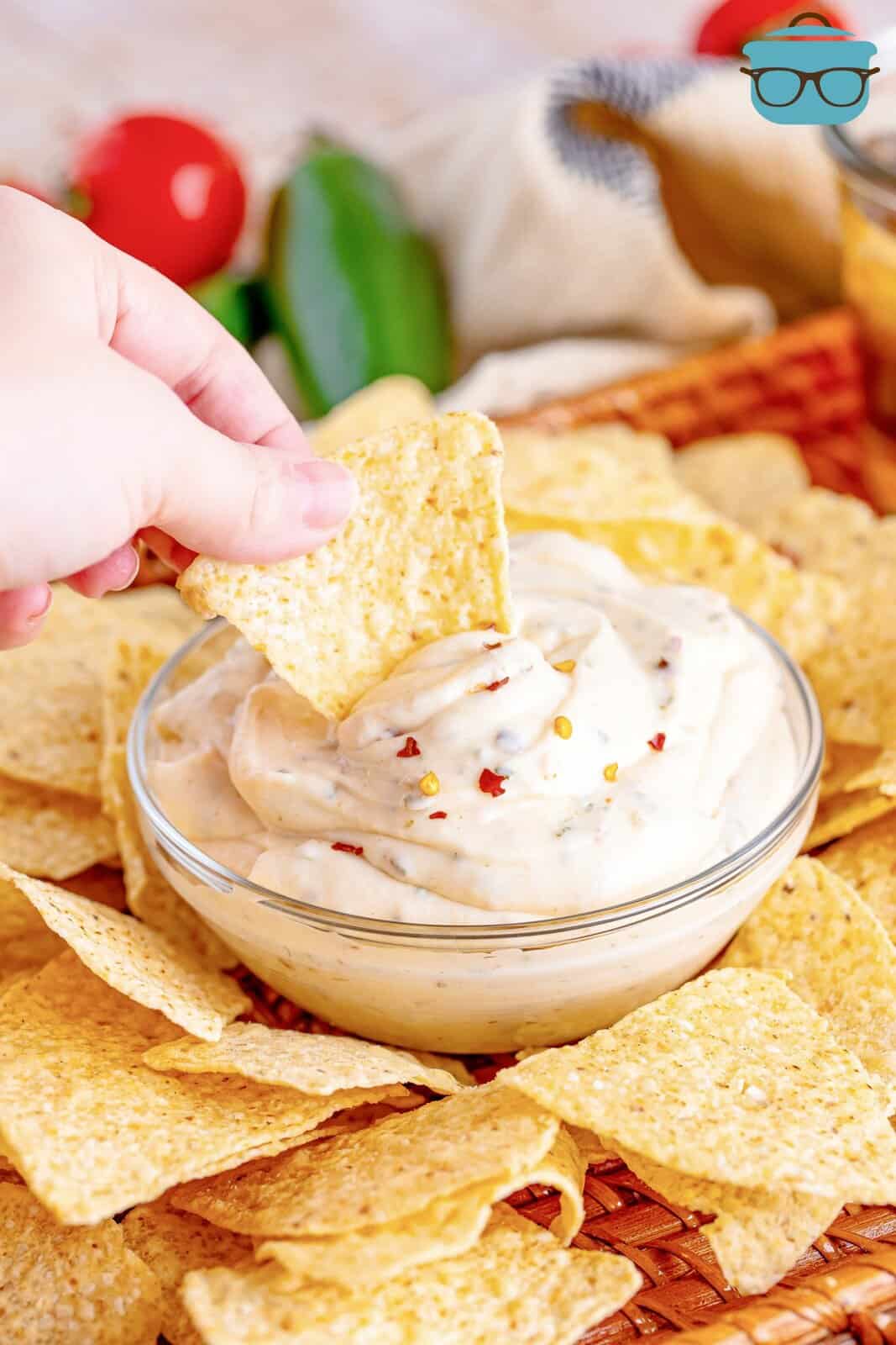 a hand holding a tortilla chip and dipping it into white sauce in a small clear bowl. 