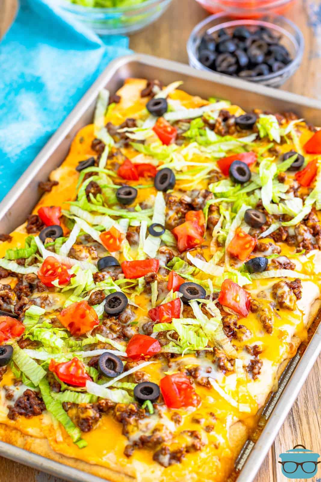 Taco pizza shown in baking sheet being topped with shredded lettuce, diced tomatoes and sliced olives. 