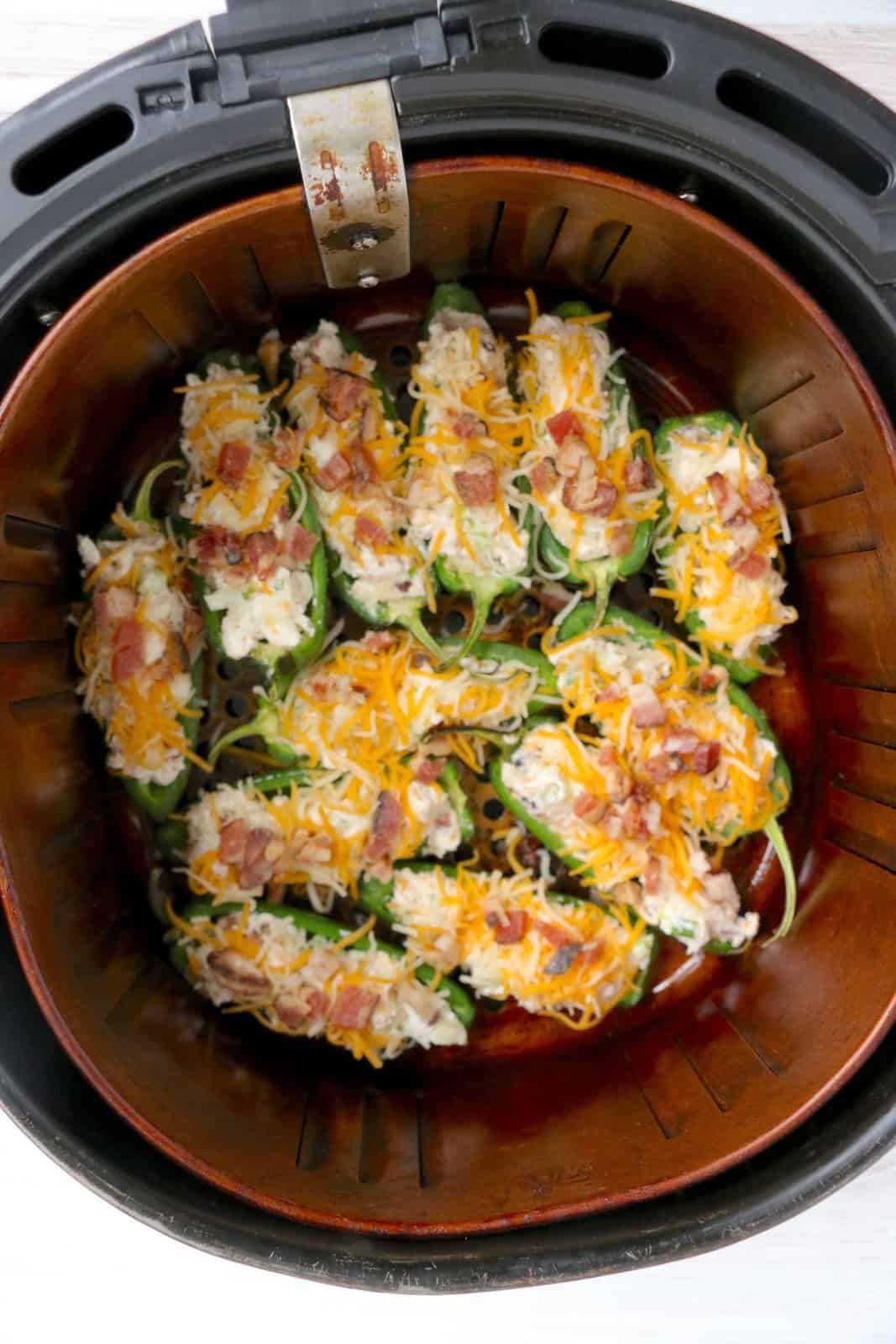 stuffed jalapenos topped with shredded cheese and chopped bacon in an air fryer basket. 