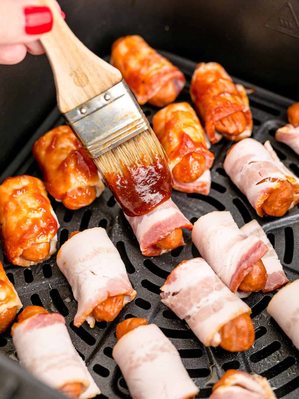 a pastry brush shown spreading barbecue sauce on top of the bacon wrapped sausages. 