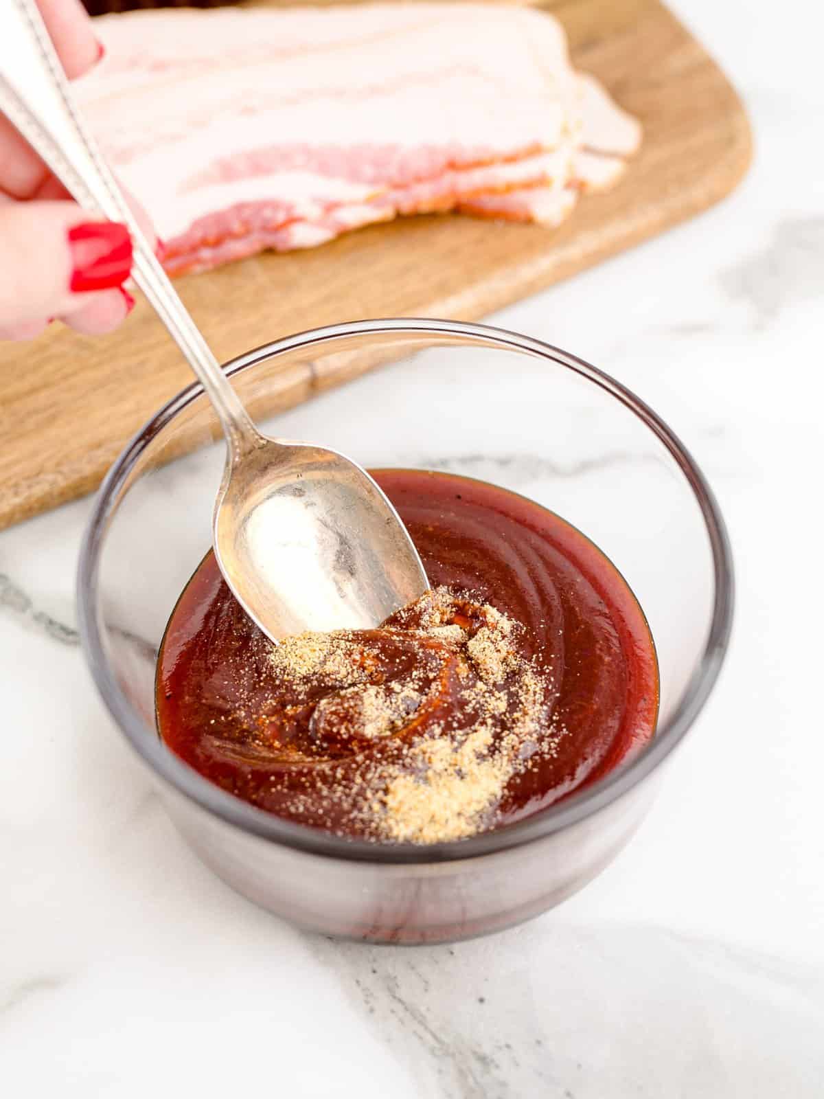 a spoon shown mixing together barbecue sauce and garlic powder in a small clear bowl.