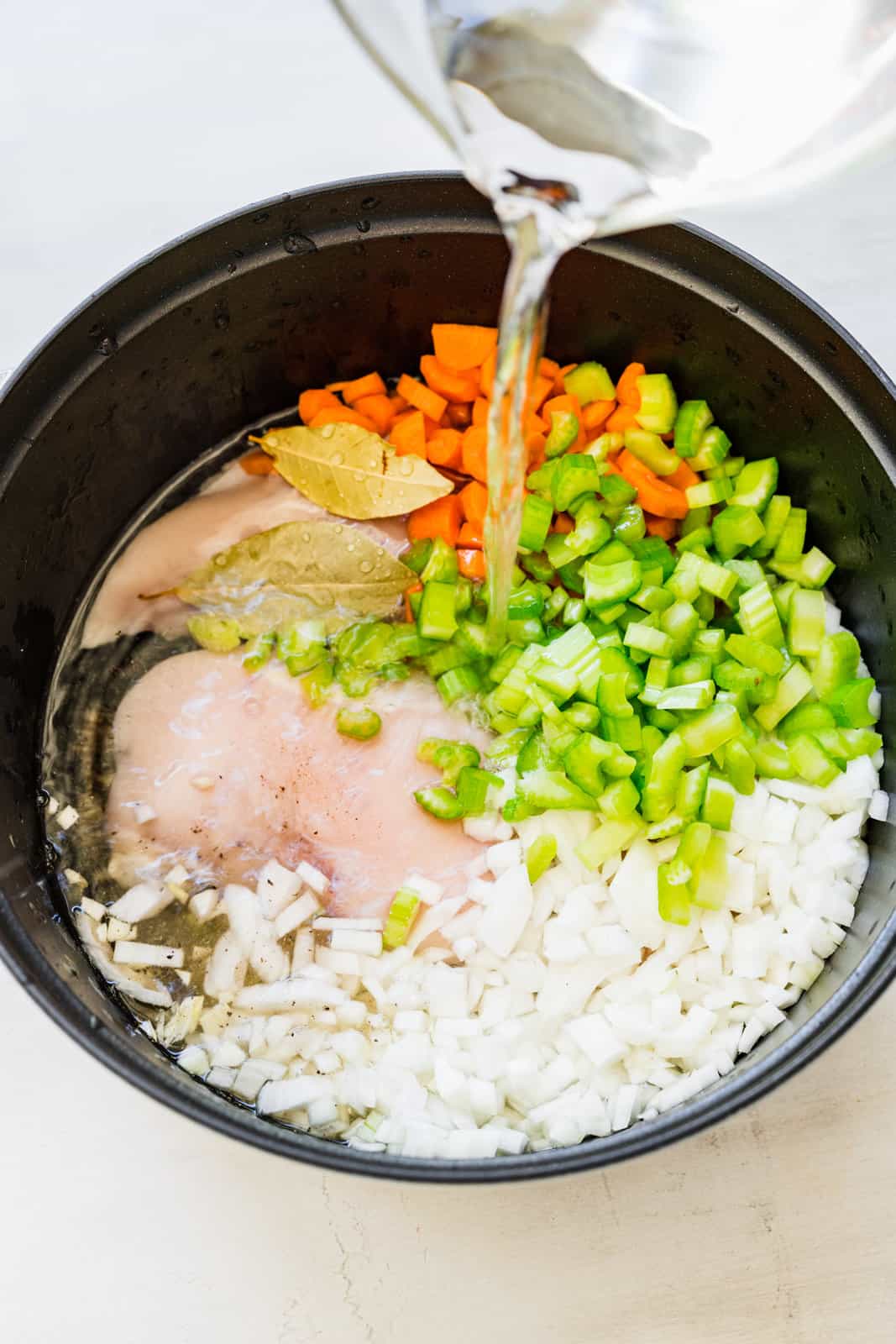 water being poured into a large pot filled with chicken breasts, diced celery, diced onion, diced carrots and seasoning. 
