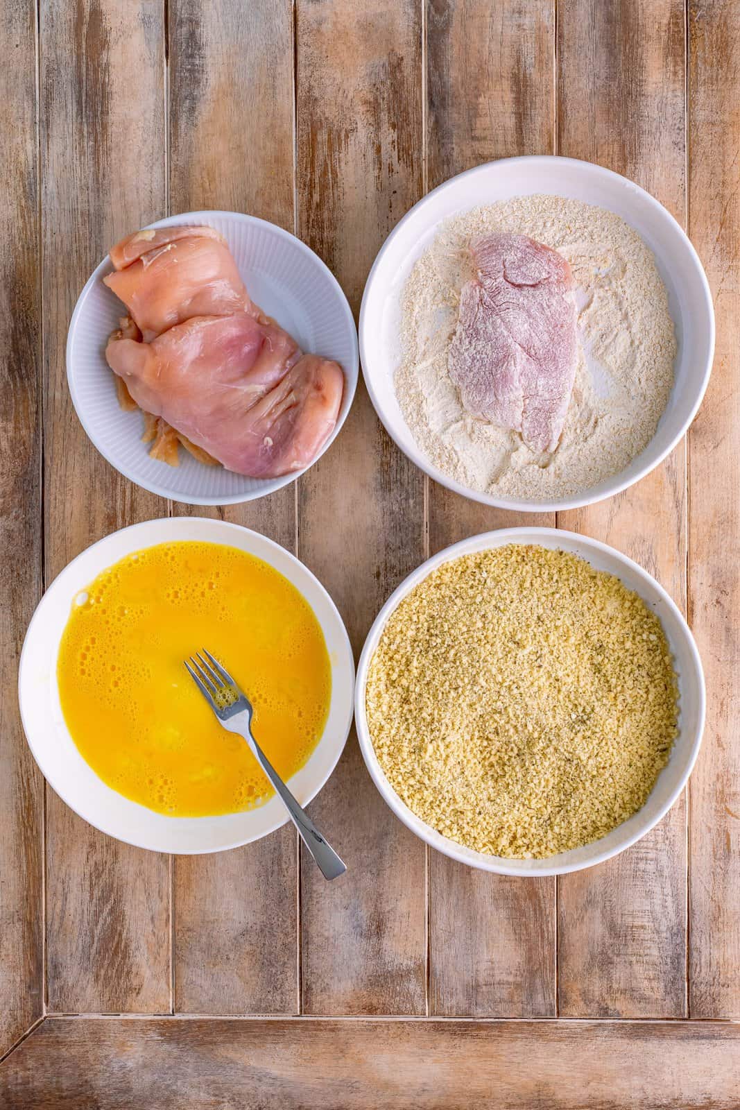 Four bowls- one with chicken, one with a piece of chicken in the flour mixture, another bowl with panko mixture and a final bowl with egg mixture. 
