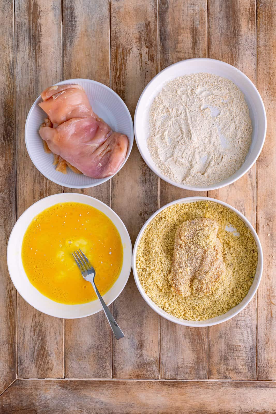 Chicken in a bowl, a bol with egg wash, a bowl with flour mixture, and a final bowl with chicken in the panko breadcrumbs.