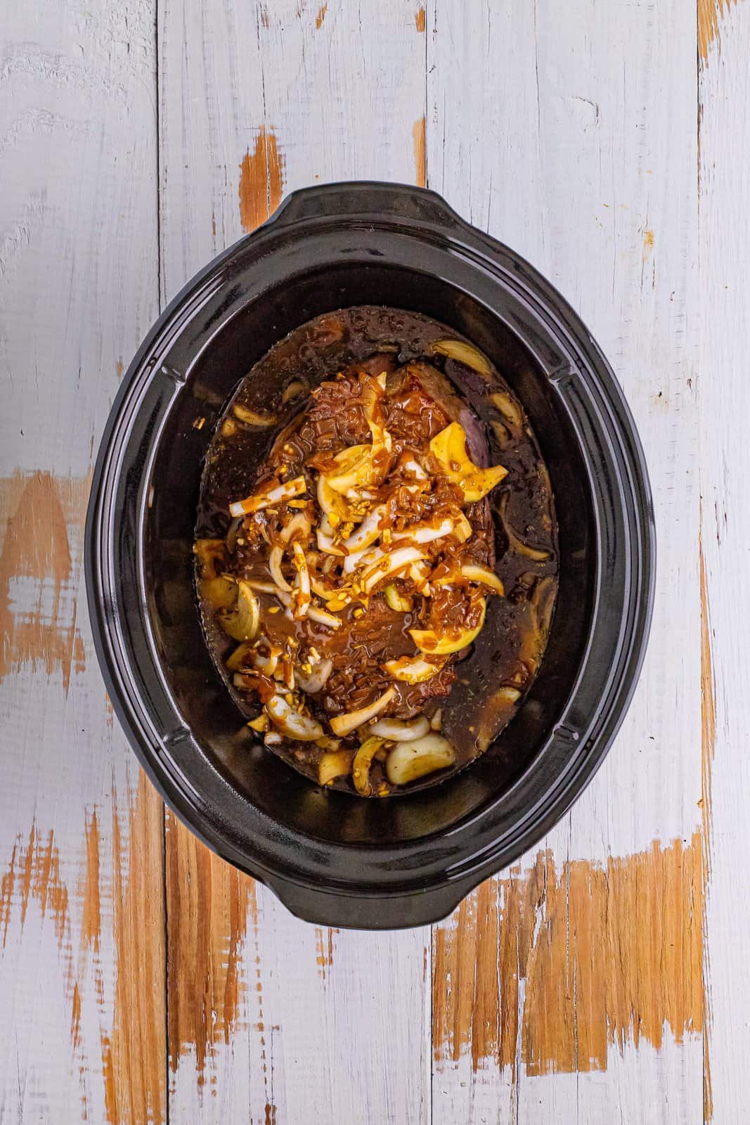 Looking down on a slow cooker with pot roast, condensed soups, onions, and herbs and spices.