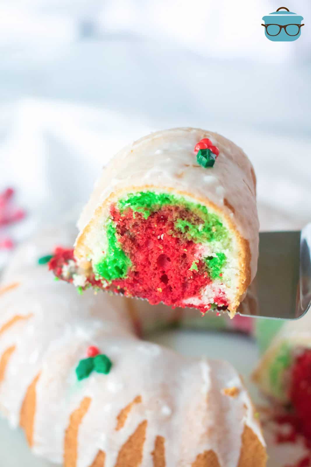 closeup photo of a spatula holding up a slice of colorful Christmas bundt cake over the cake. 