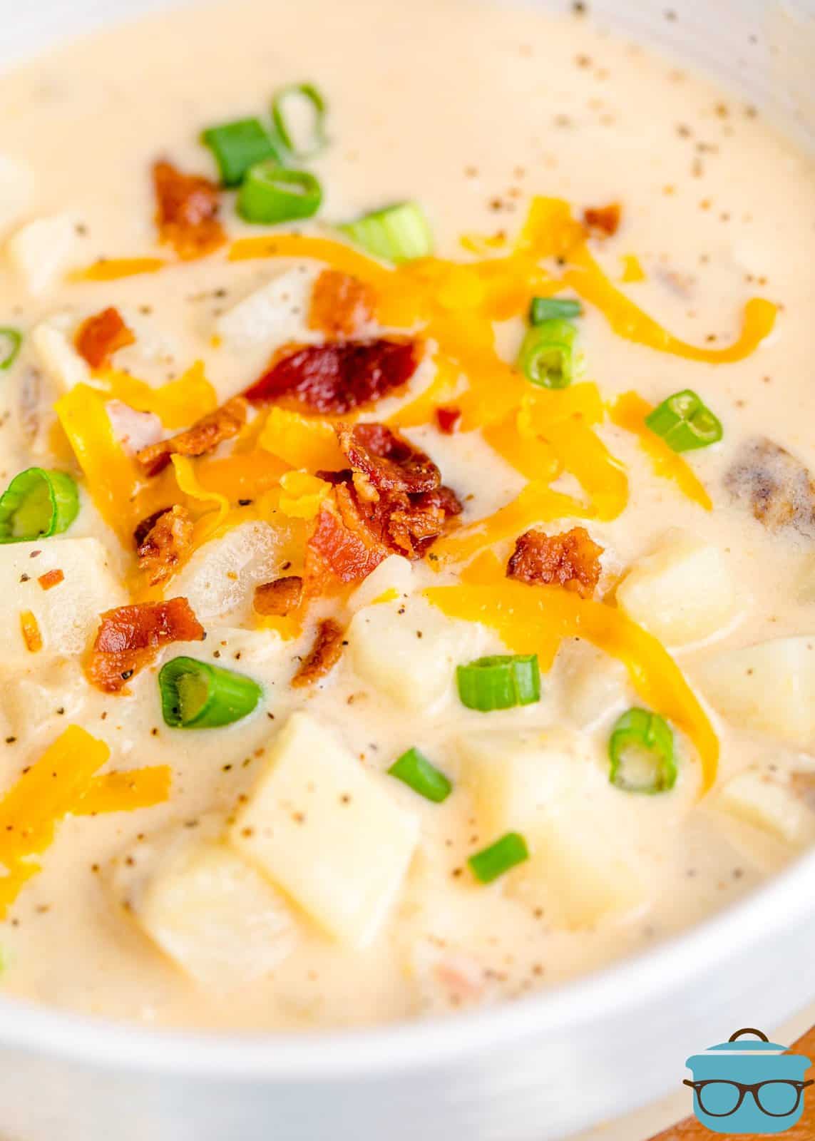 close up photo of potato soup in a bowl topped with crumbled bacon, shredded cheddar cheese and sliced green onions. 