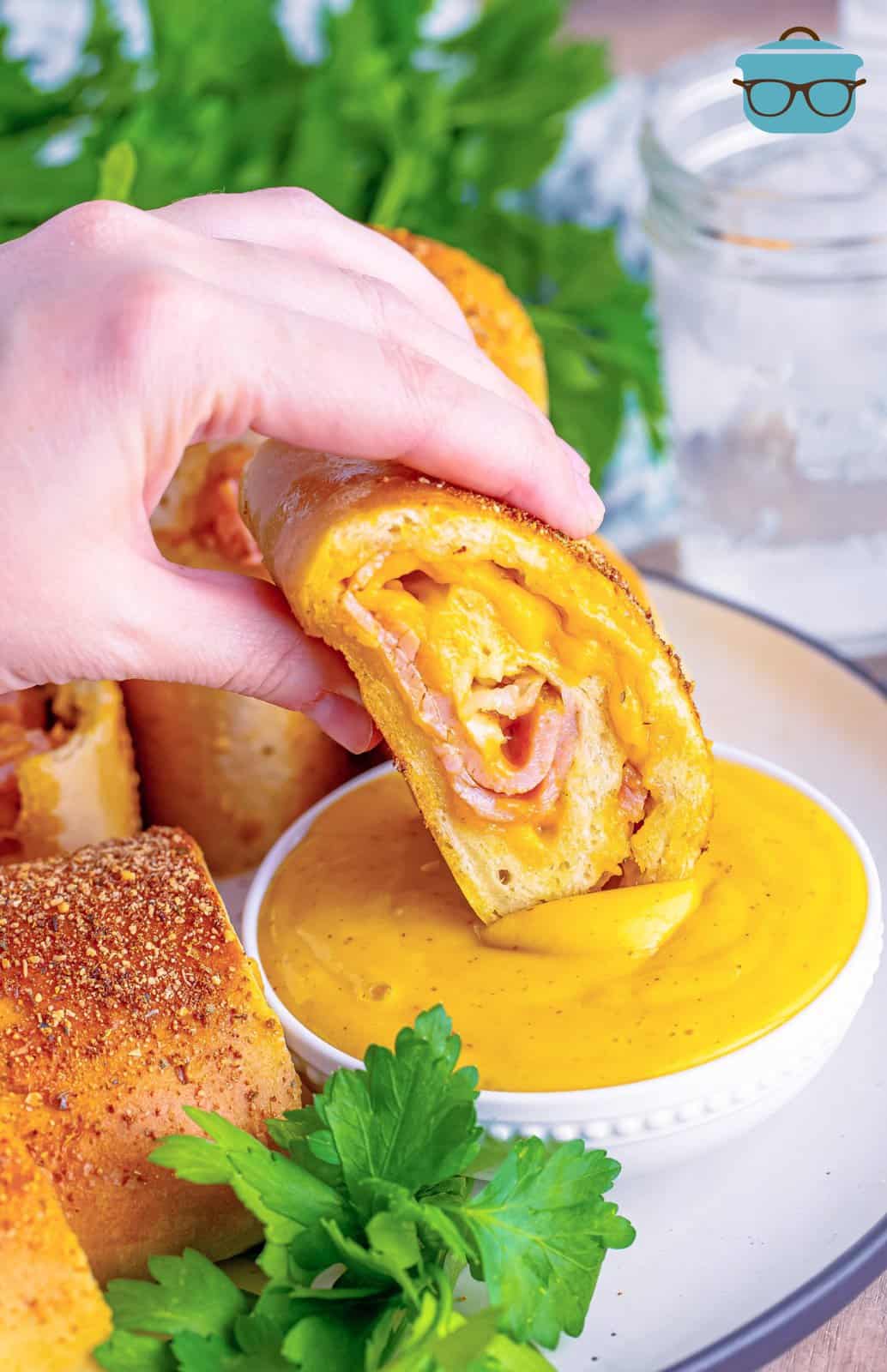 a hand holding a slice of ham and cheese Stromboli and dipping it into honey mustard sauce. 