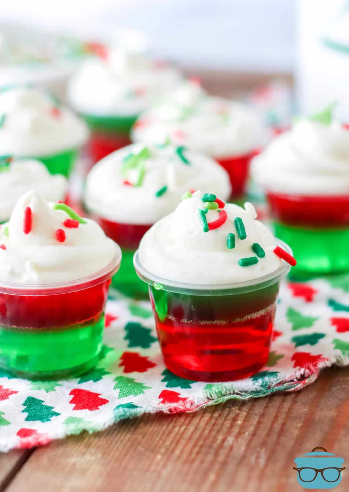 Holiday Jell-O shots on Christmas linen topped with whipped topping and sprinkles.