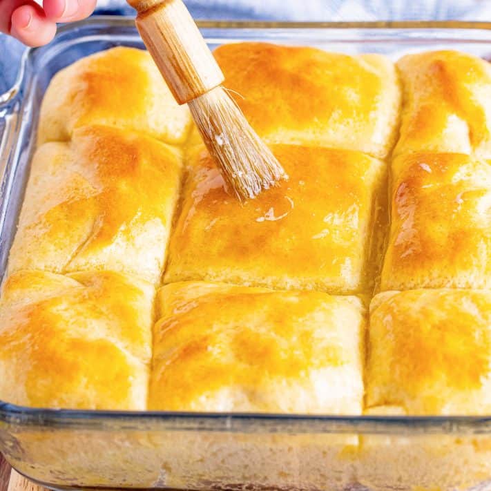 A glass pan of yeast rolls and a pastry brush brushing butter on top.