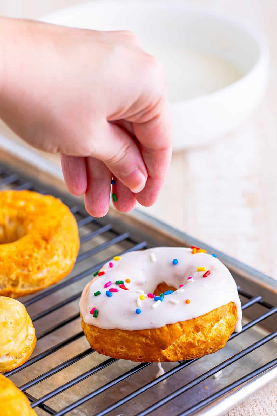 A hand sprinkling sprinkles on a vanilla glazed donut.