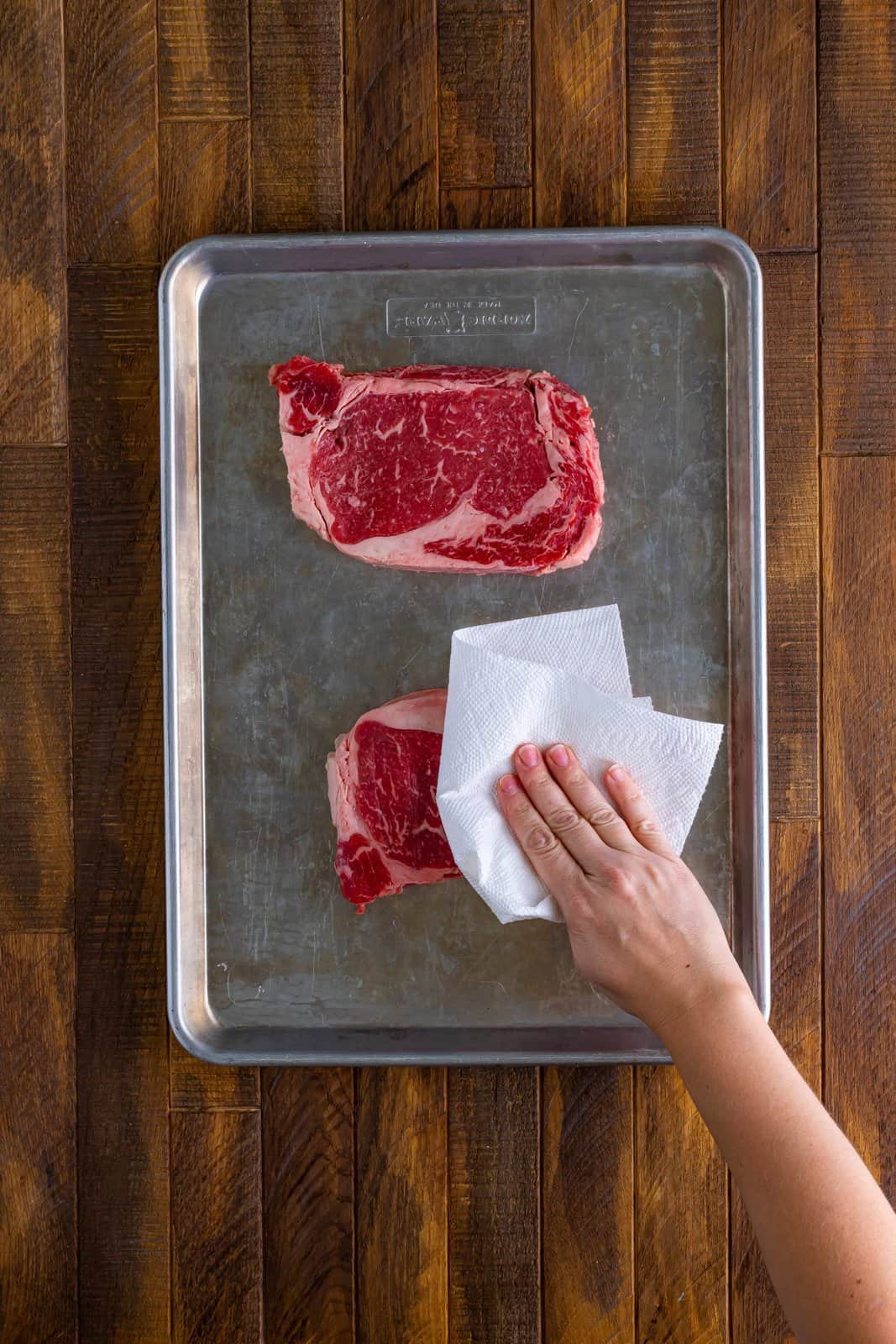 A sheet pan with two raw steaks and a hand patting them dry with a paper towel.